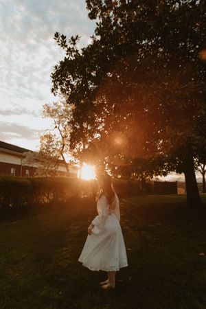 A figure in a white dress twirls in a garden at sunset with dramatic lens flare and tree silhouettes.
