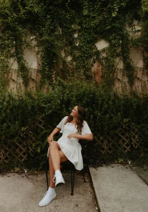 Casual portrait in white dress and sneakers against ivy-covered wall.