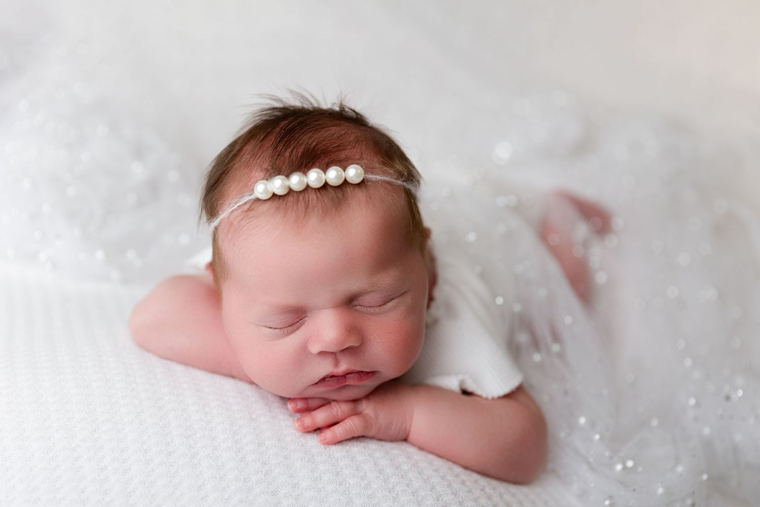 Peaceful newborn sleeping on white textured blanket wearing pearl headband.