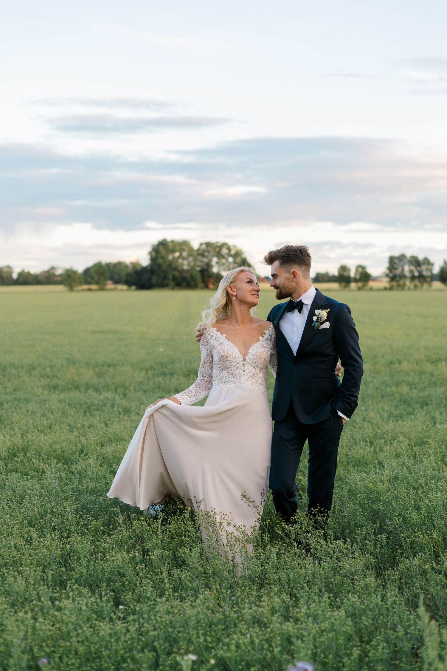 A wedding couple poses together in a scenic green field at sunset, with her lace dress flowing in the breeze.