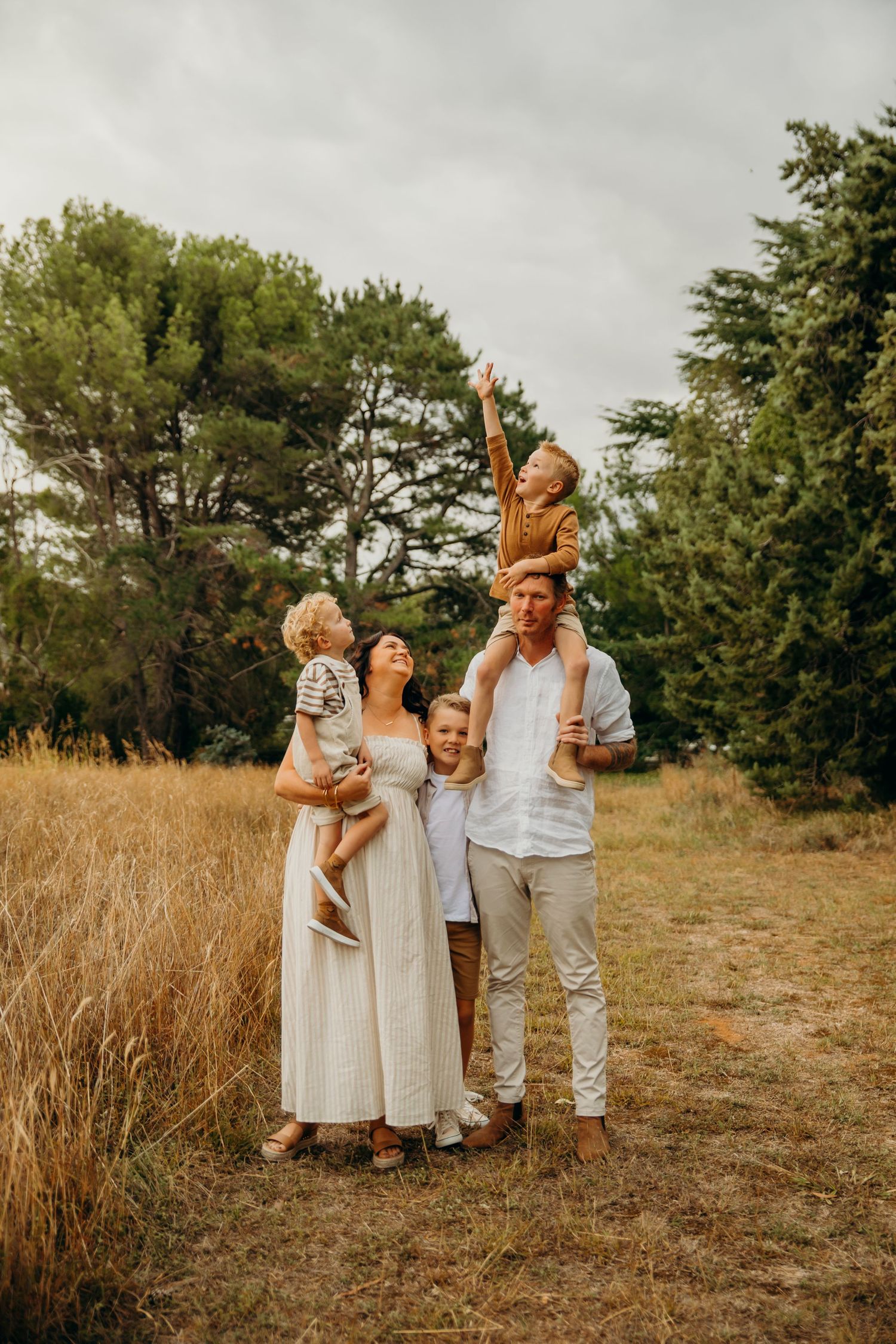 Family of five poses in golden field with trees, children laughing as boy reaches skyward.
