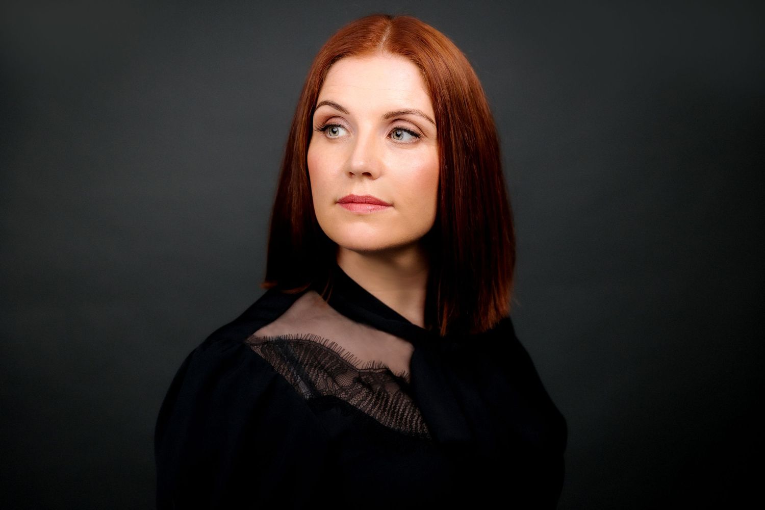Headshot of woman looking off camera against dark background with auburn hair styled straight frames features in dramatic lighting effect.