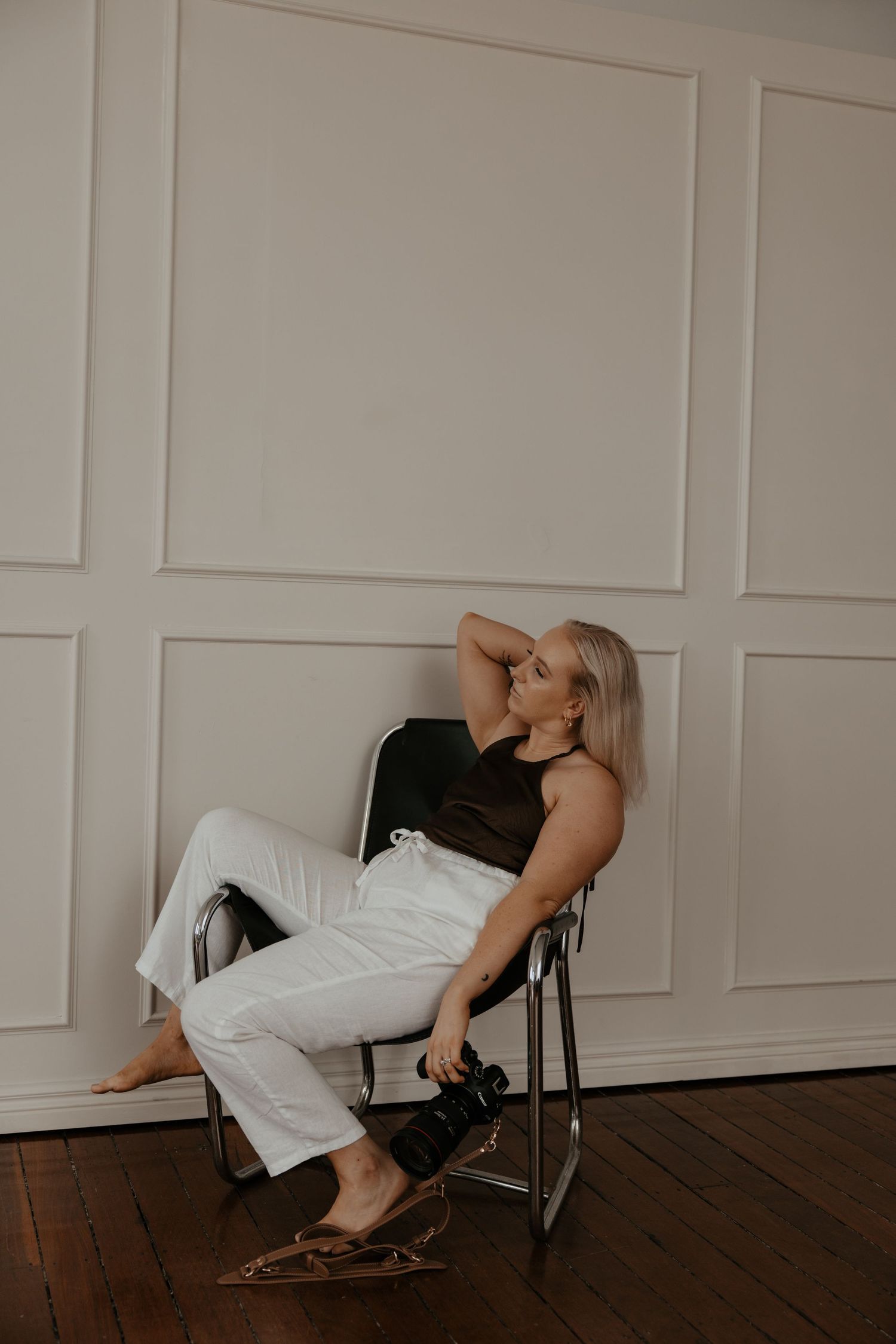 Woman in white pants and black top lounges in a chair against a white paneled wall.