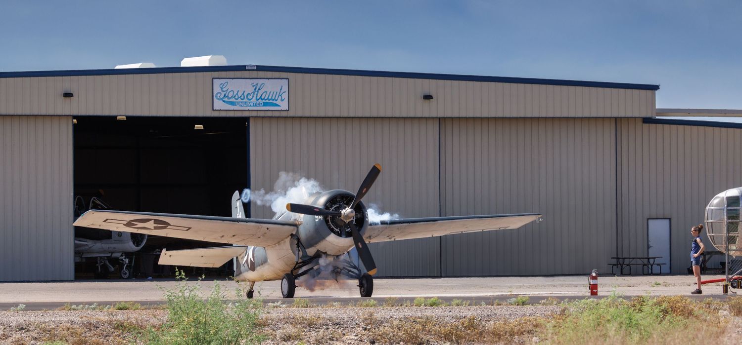 Vintage WWII propeller aircraft parked on tarmac in front of a large metal hangar at a small airfield.