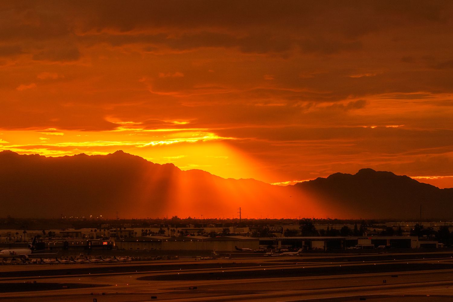 Dramatic orange sunset with golden sun rays bursting over mountain silhouettes above an airport runway.