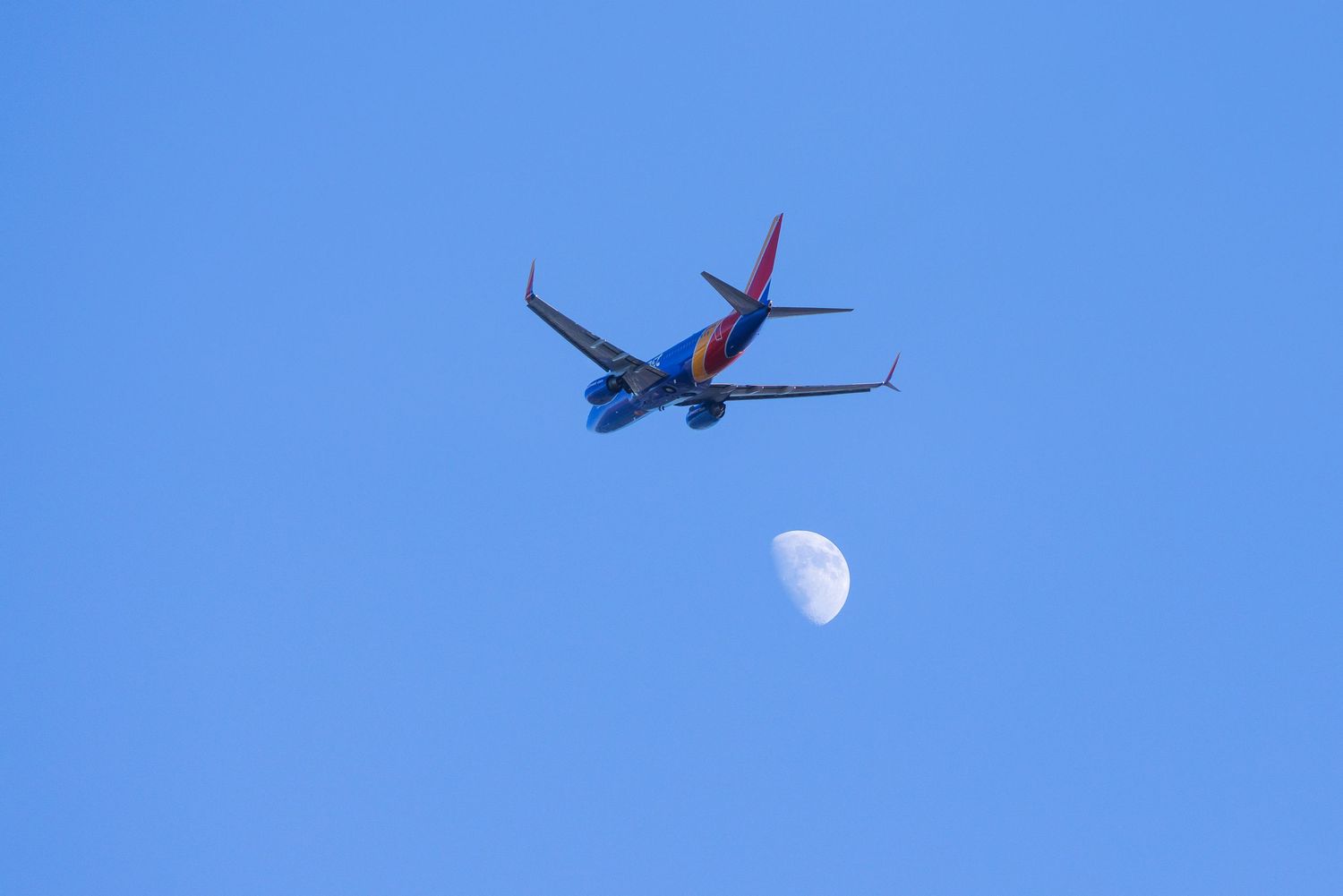 Southwest Airlines plane flies through clear blue sky with a half moon visible below it.
