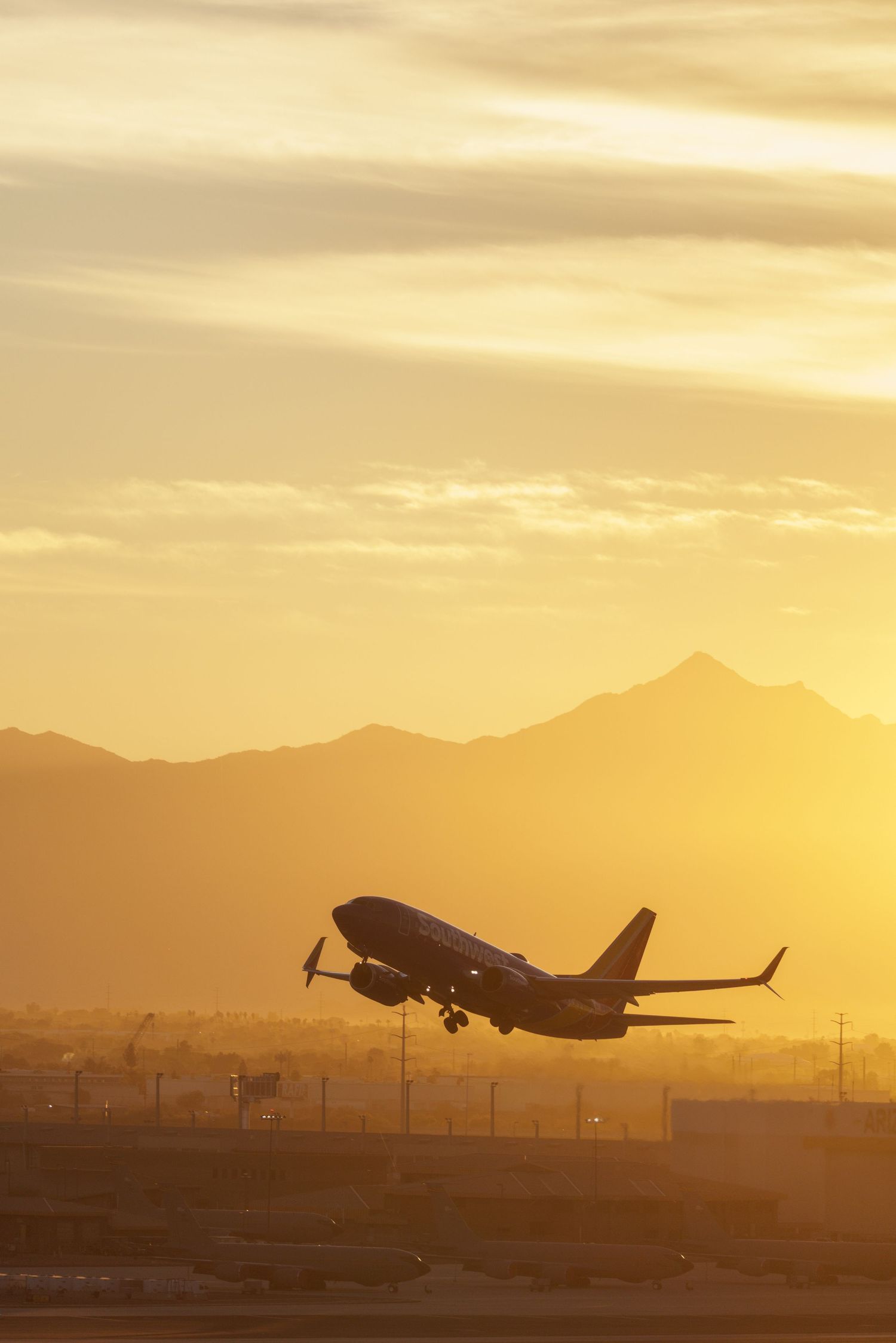 Airplane silhouette taking off at sunset with golden sky and mountain range backdrop at a busy airport.