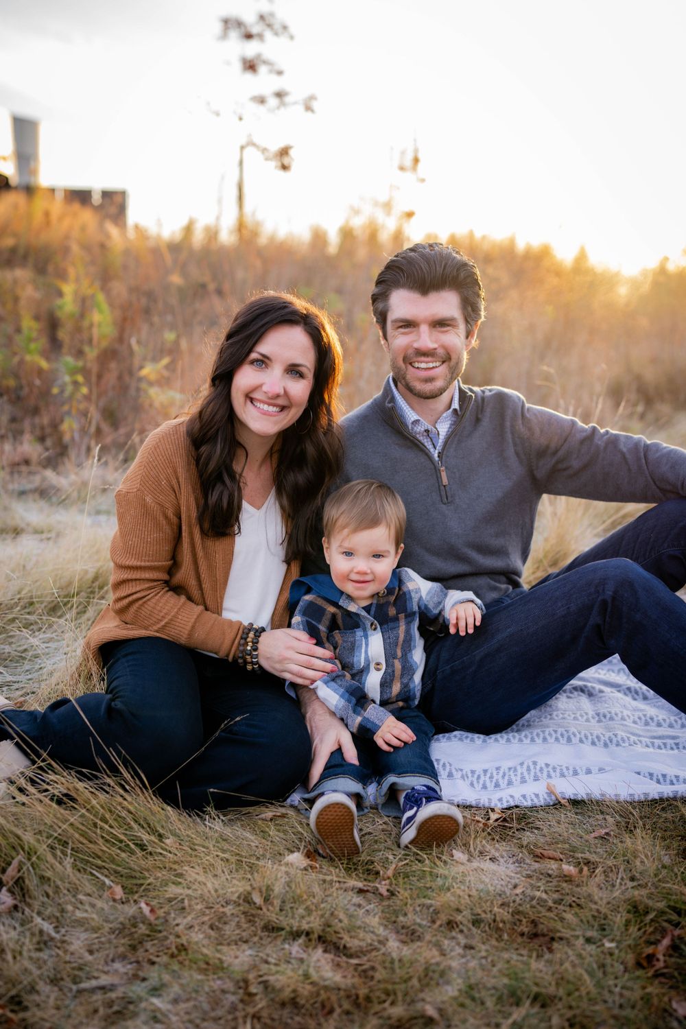 Smiling family with toddler sitting in golden field at sunset.