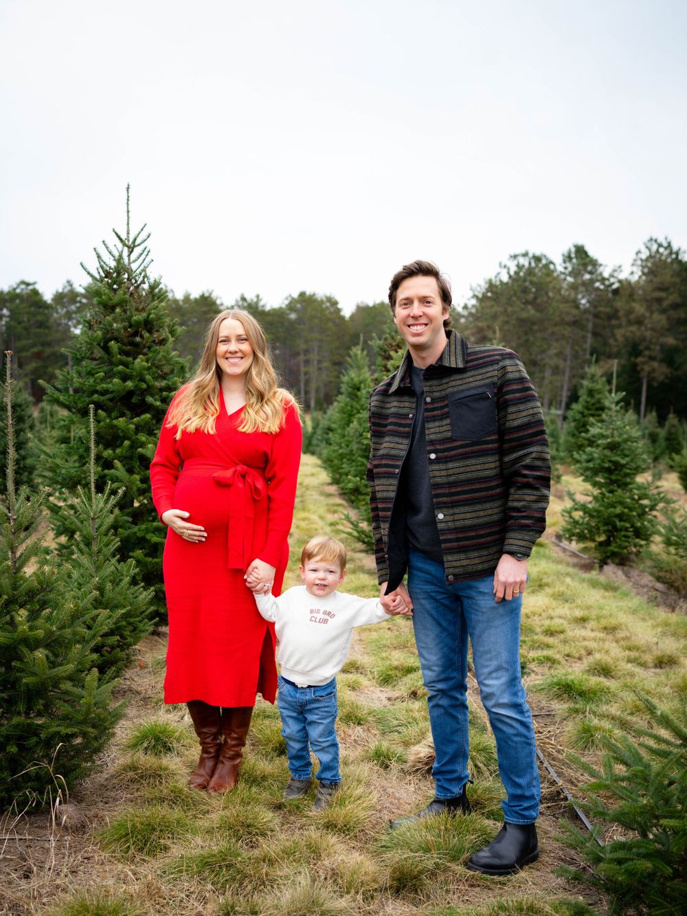 Pregnant woman in red dress with family at Christmas tree farm.