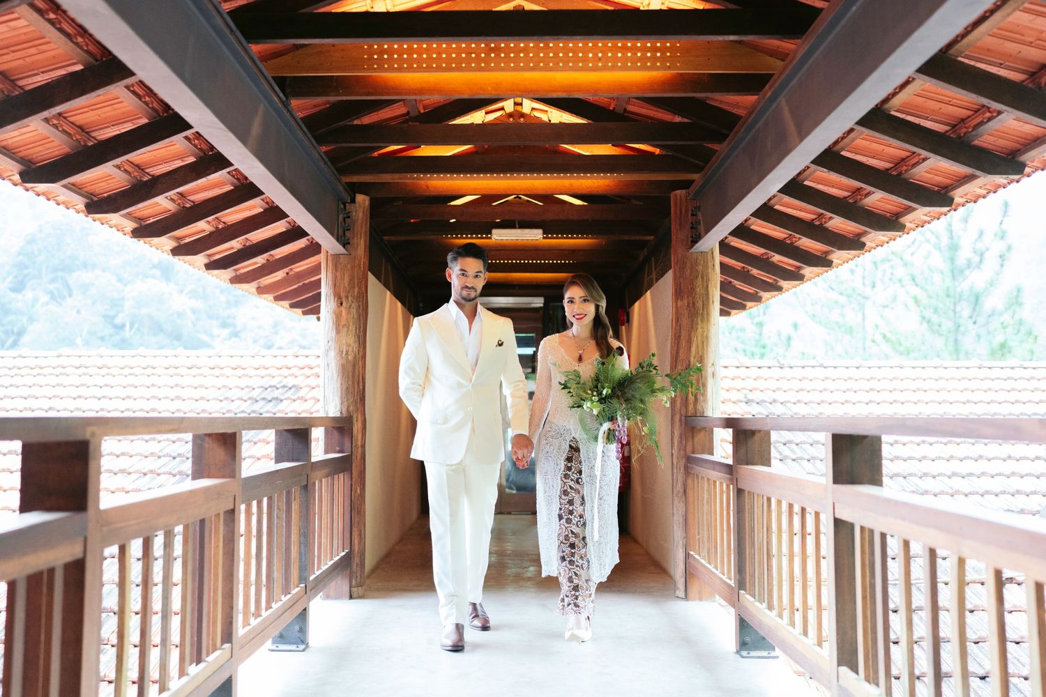 Elegant wedding couple in white attire walking hand in hand through a wooden covered bridge.