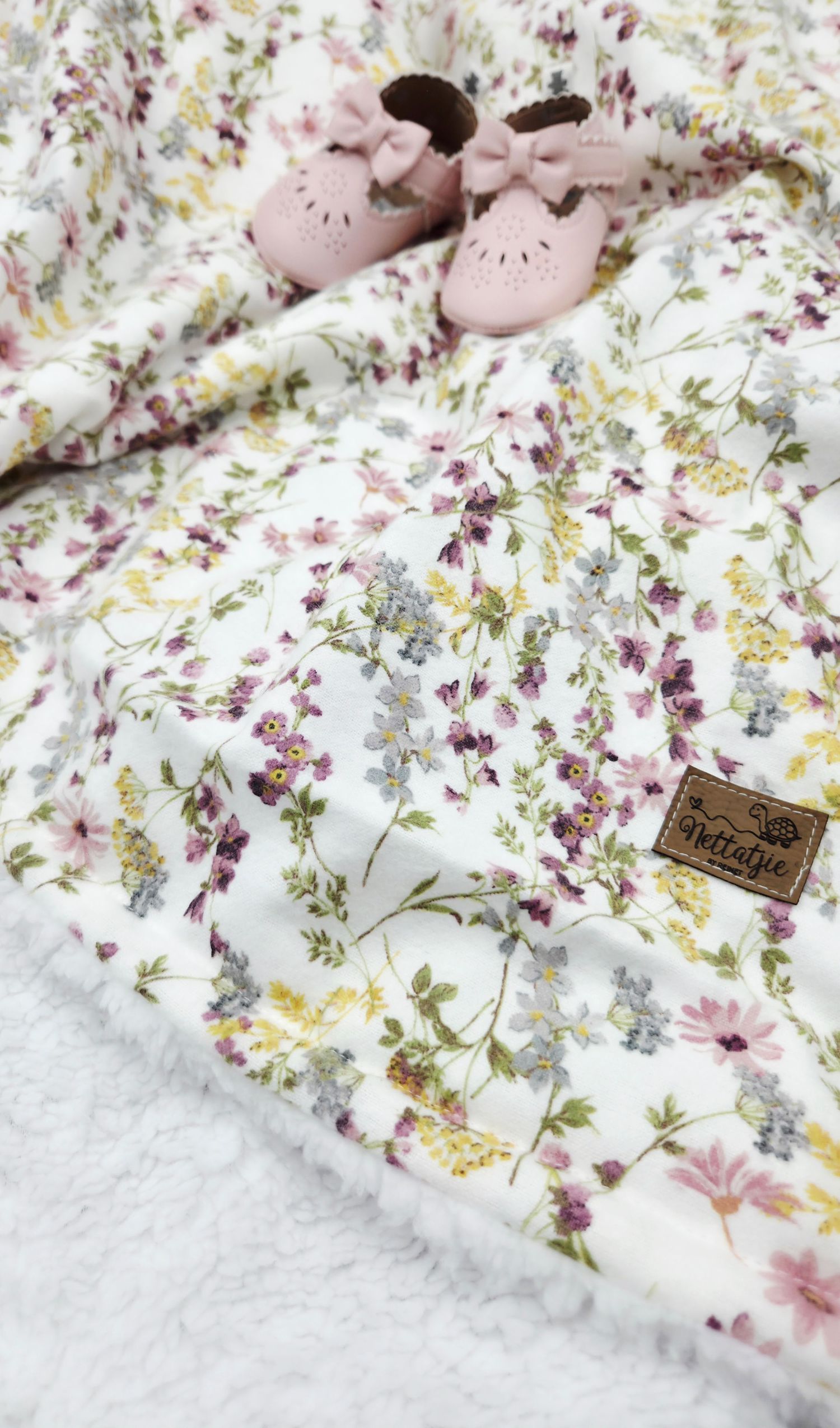 Soft floral baby blanket with pink and purple wildflowers, paired with pink bow baby shoes on white sherpa fabric.
