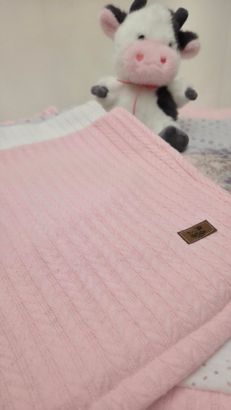 Pink cable-knit baby blanket with leather logo patch, displayed with a cute black and white cow stuffed animal toy.