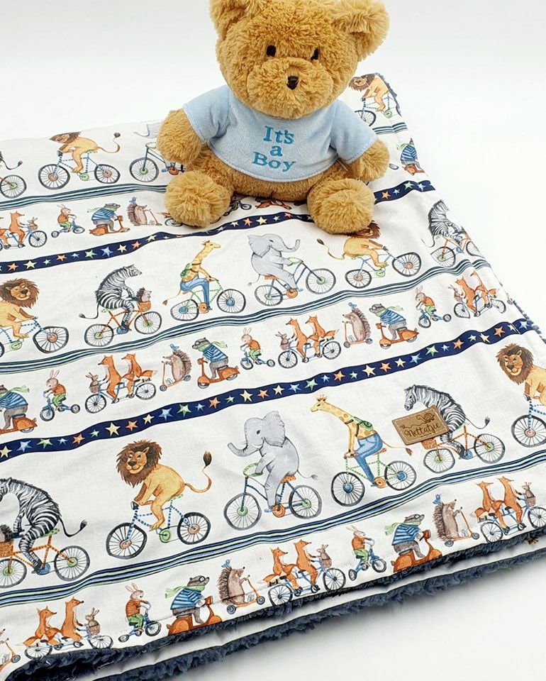 A plush teddy bear in a blue 'It's a Boy' shirt sits on a baby blanket with whimsical animal bicycle pattern design.