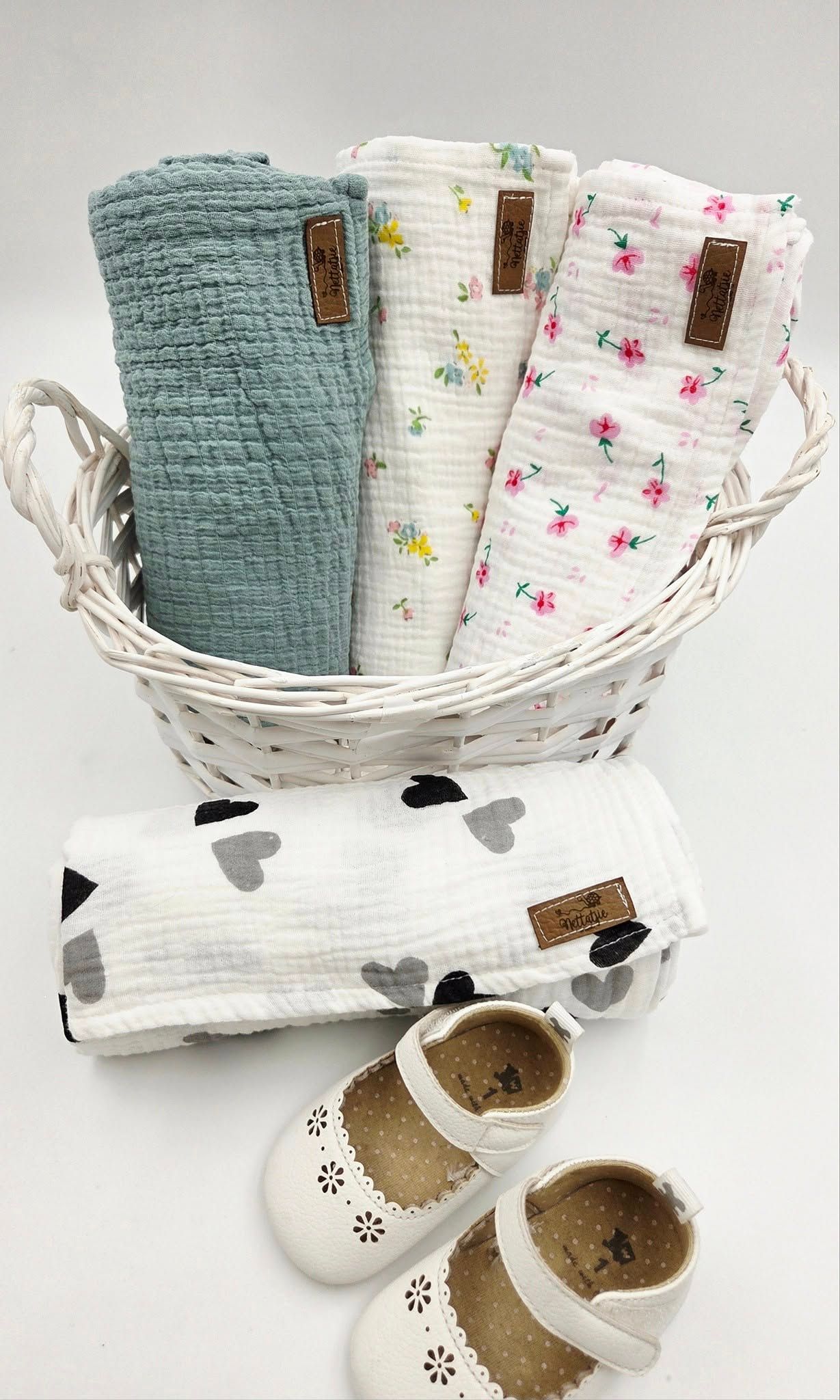 White wicker basket containing folded baby blankets and white infant shoes.