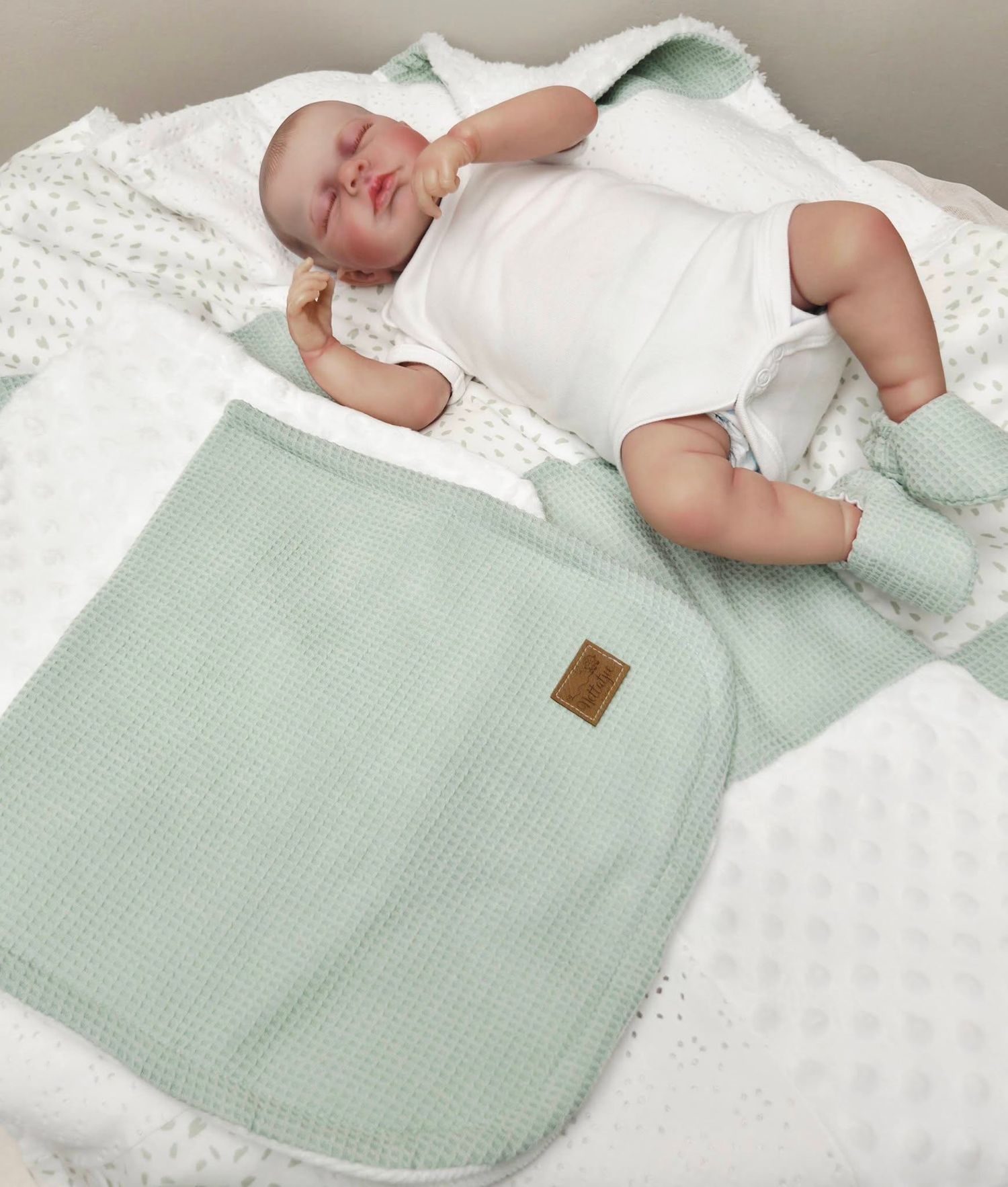 Newborn baby resting on a soft white blanket with a mint green waffle-knit sleep sack and matching booties.
