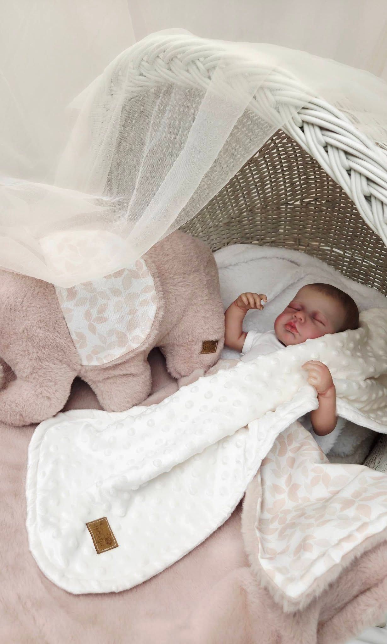 Sleeping reborn baby doll in a white wicker bassinet with a pink elephant plush toy and soft floral blanket.