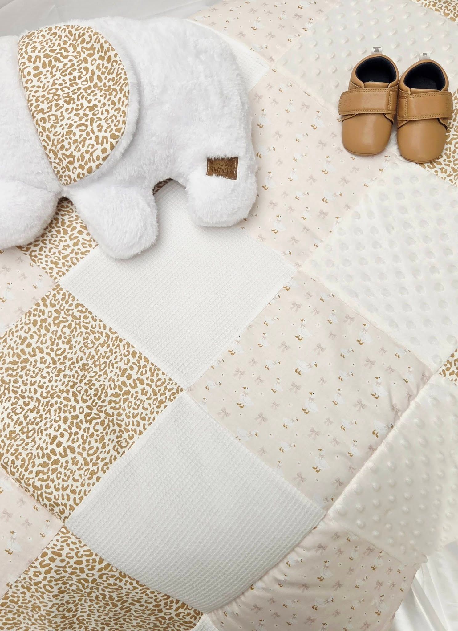 Neutral tone patchwork baby quilt with leopard print, white elephant plush toy, and tan leather baby shoes.