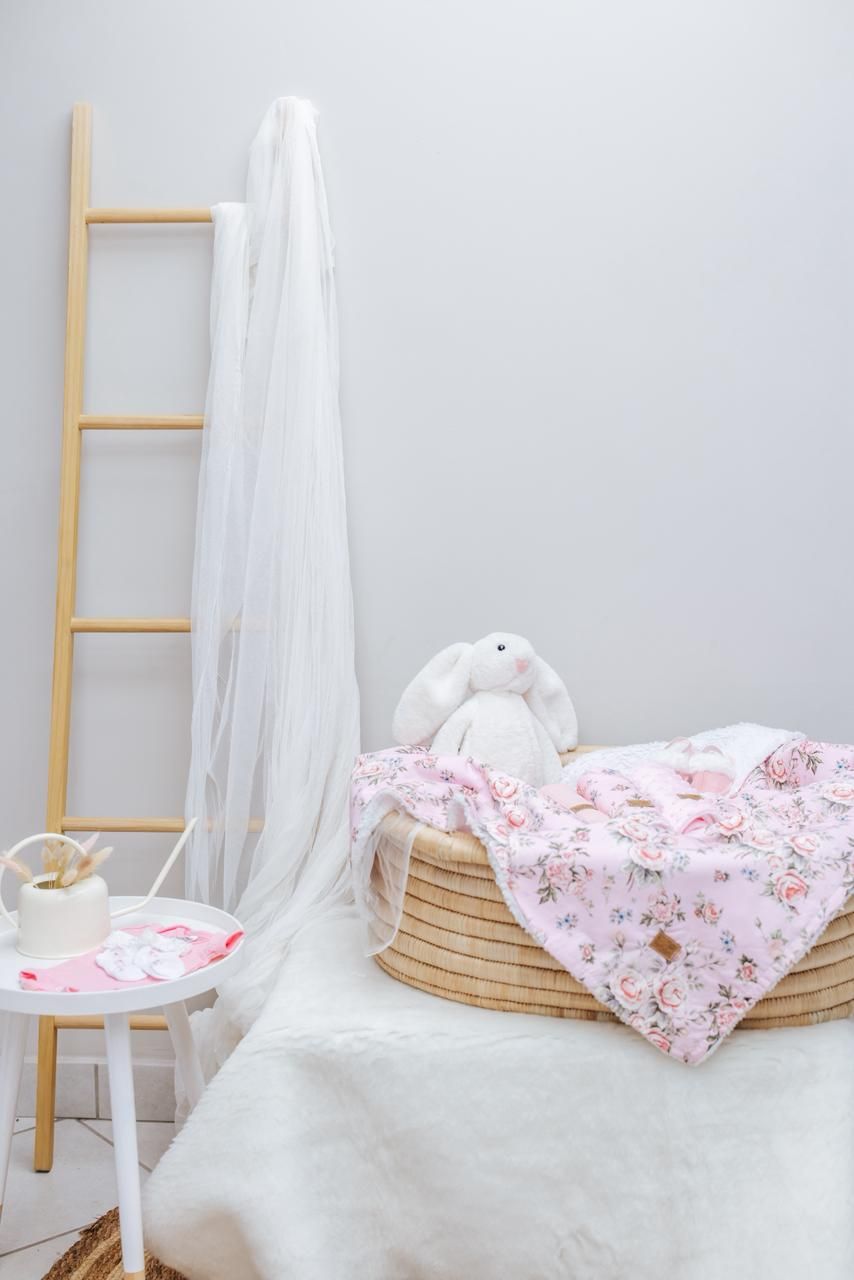 Pink floral blanket and white bunny plush arranged in woven basket beside wooden ladder and white stool.