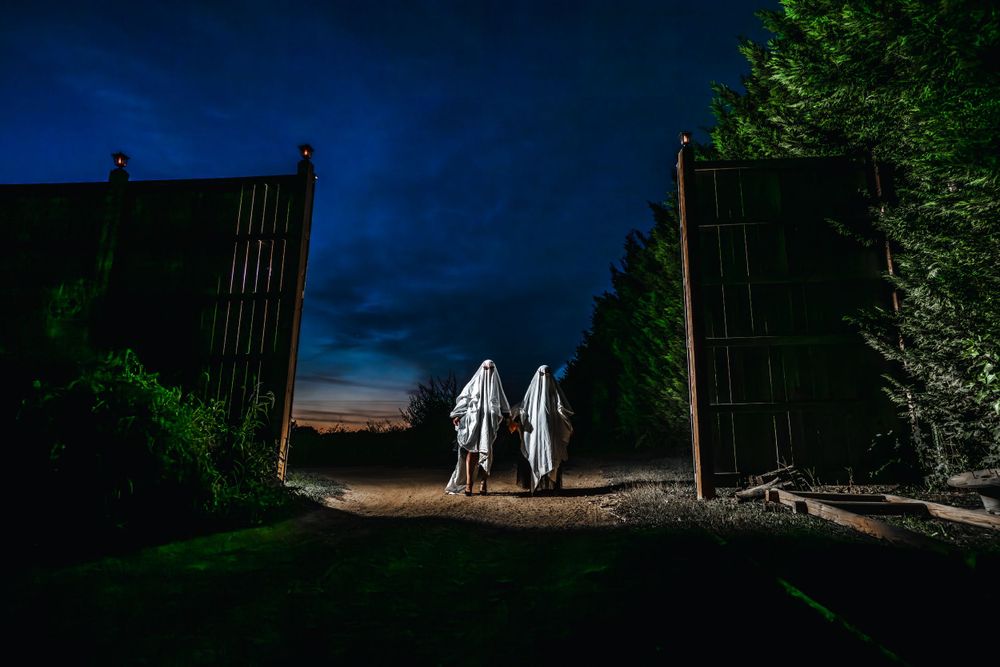Silhouettes of two figures in white ghost costumes walking through open gates at twilight.