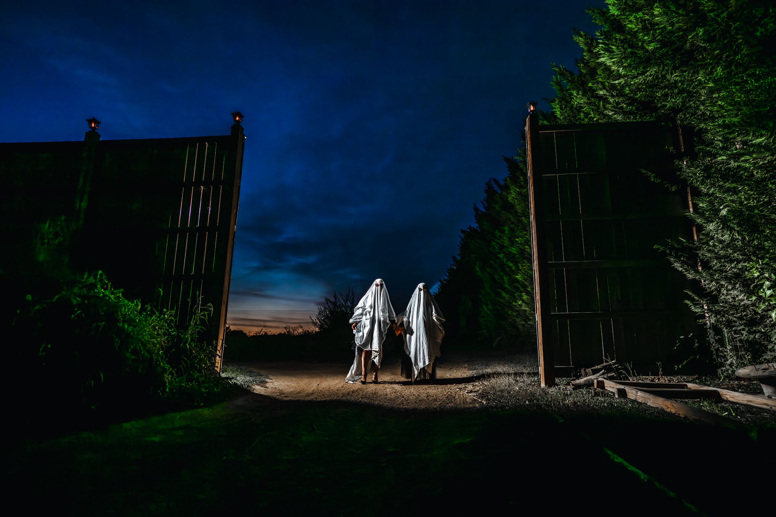 Silhouettes of two figures in white ghost costumes walking through open gates at twilight.