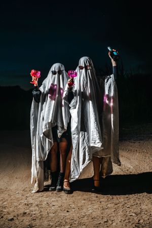 Two people in ghost costumes holding cocktails pose on dirt road at night.