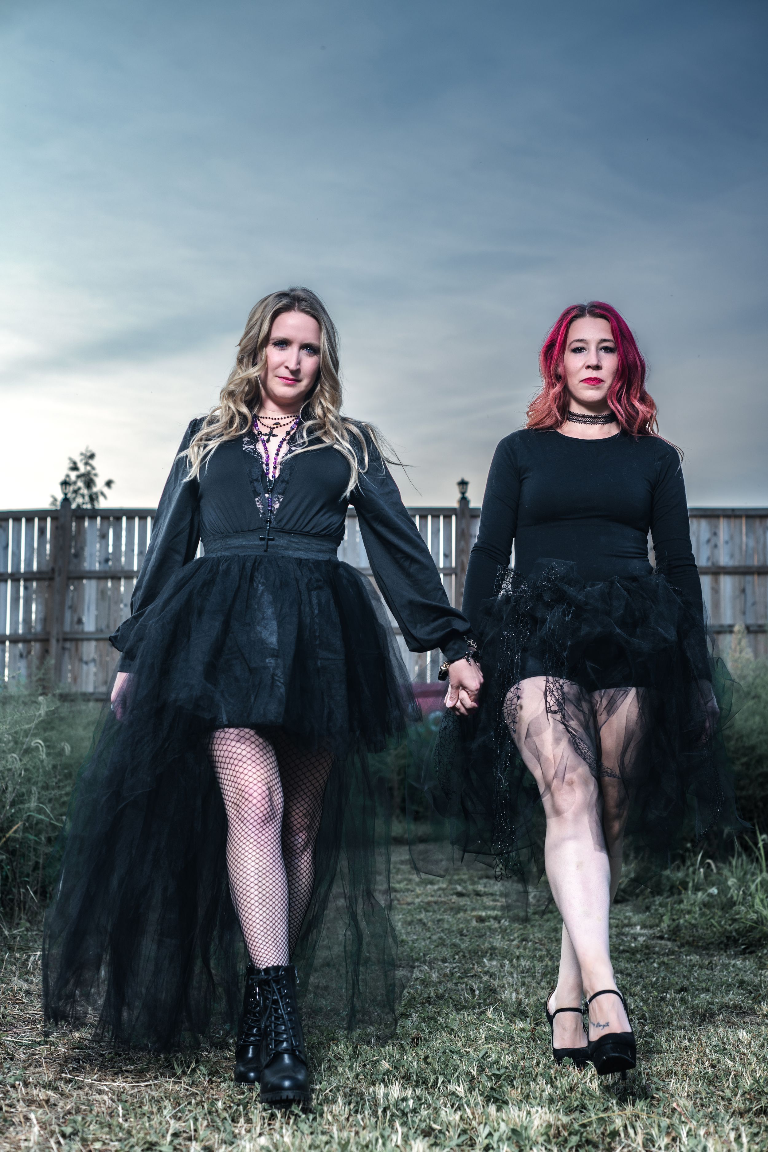 Two women in black gothic dresses and fishnet stockings pose outdoors against a moody sky.