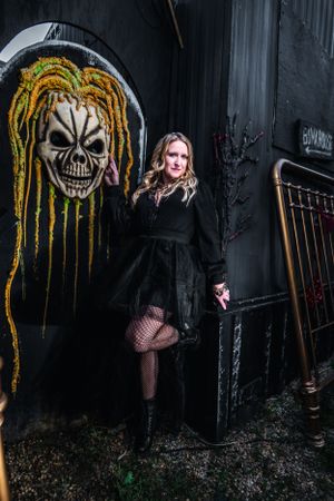 A person in gothic attire poses by street art featuring a skull with yellow dreadlocks.