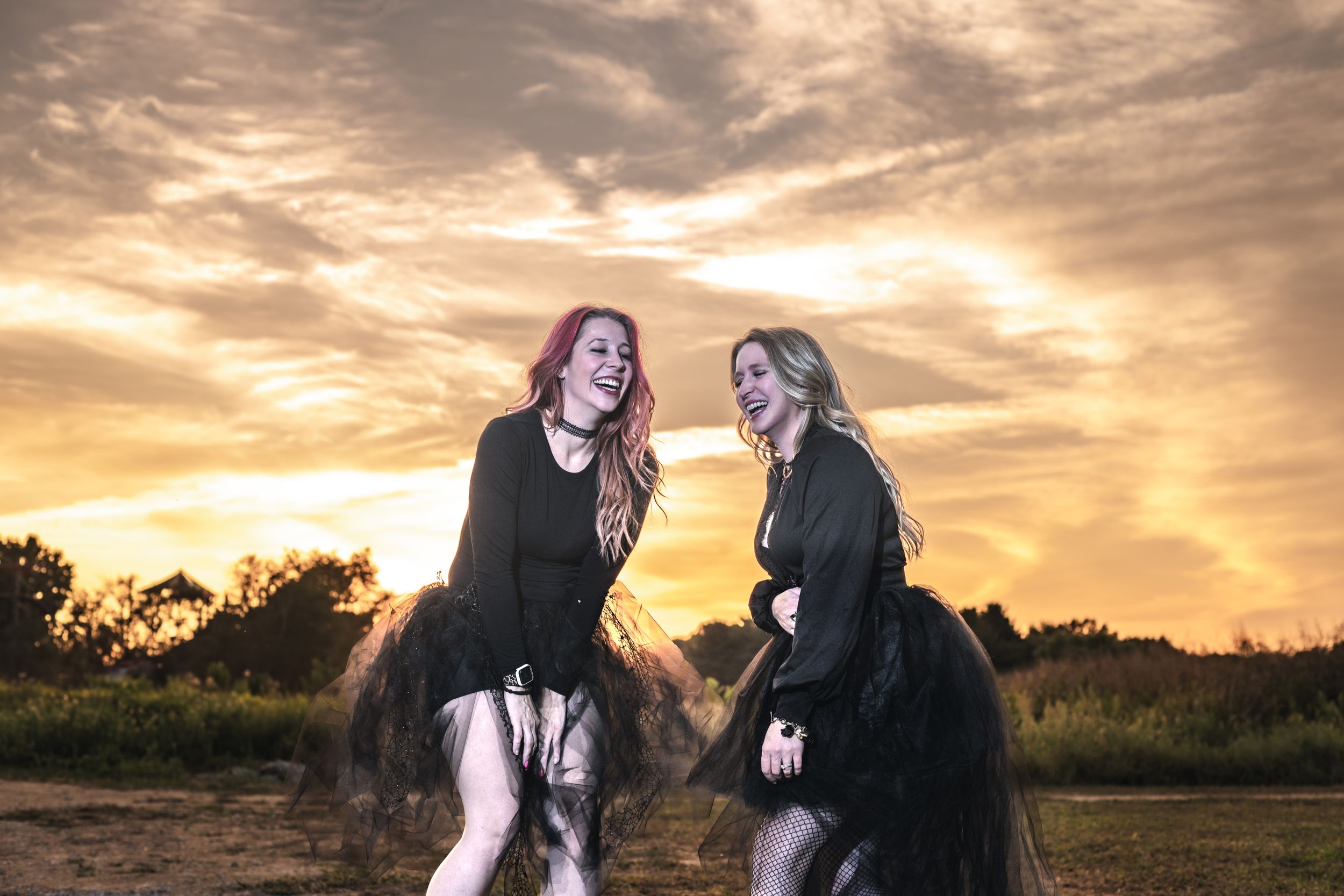 Two friends in dark dresses dance together at sunset in a field.