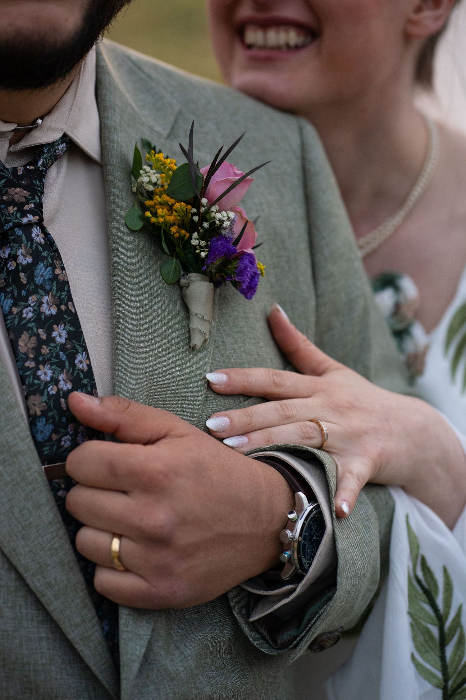 Bride smiles while embracing her groom in intimate detail photo showing boutonnière, watch, and rings