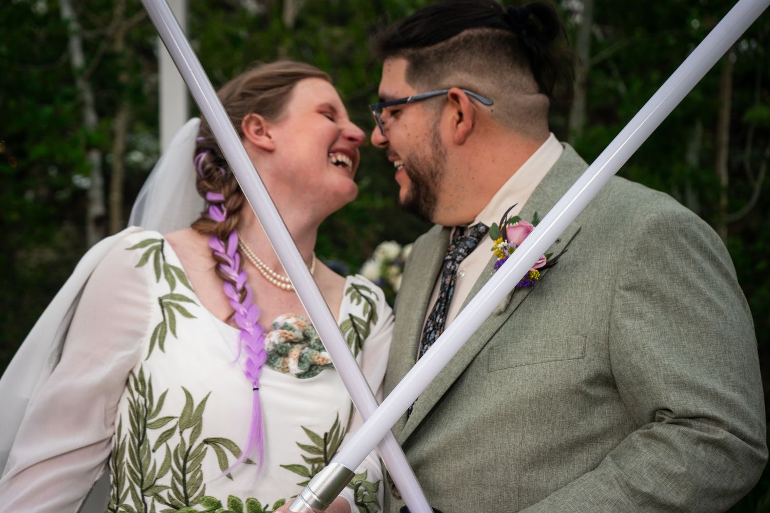 A couple with light sabers shares a romantic moment during their outdoor wedding ceremony surrounded by greenery and white decor.