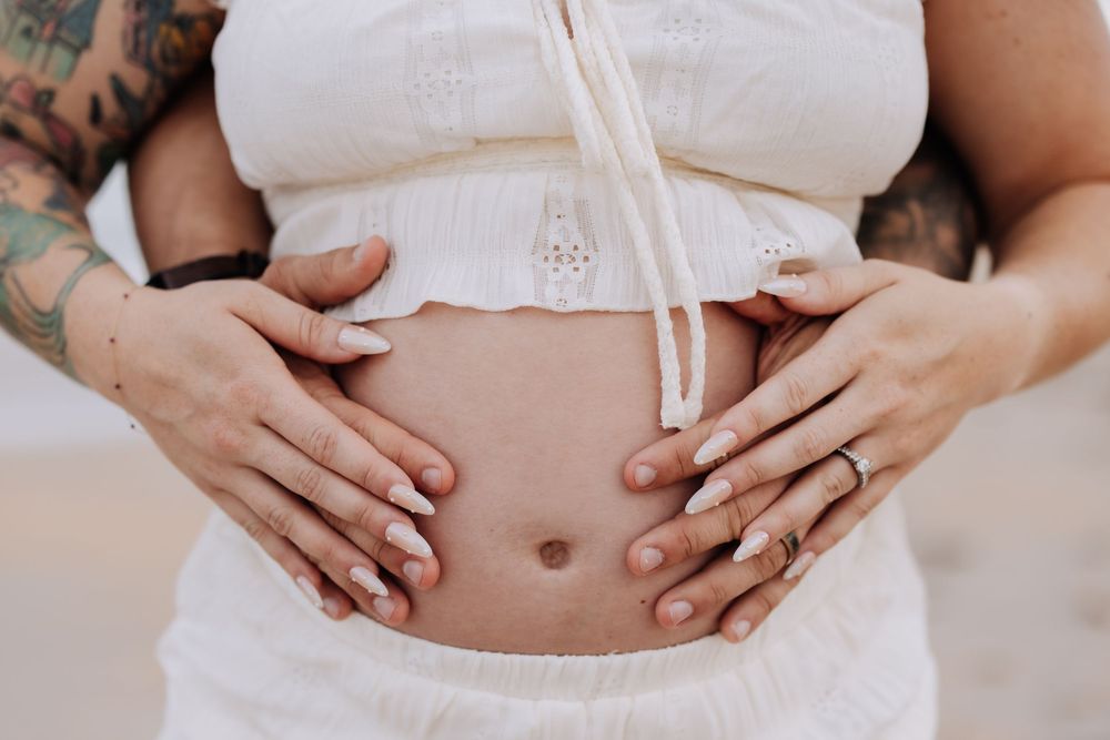 Pregnant woman in white top with tattooed partner's hands cradling baby bump outdoors.