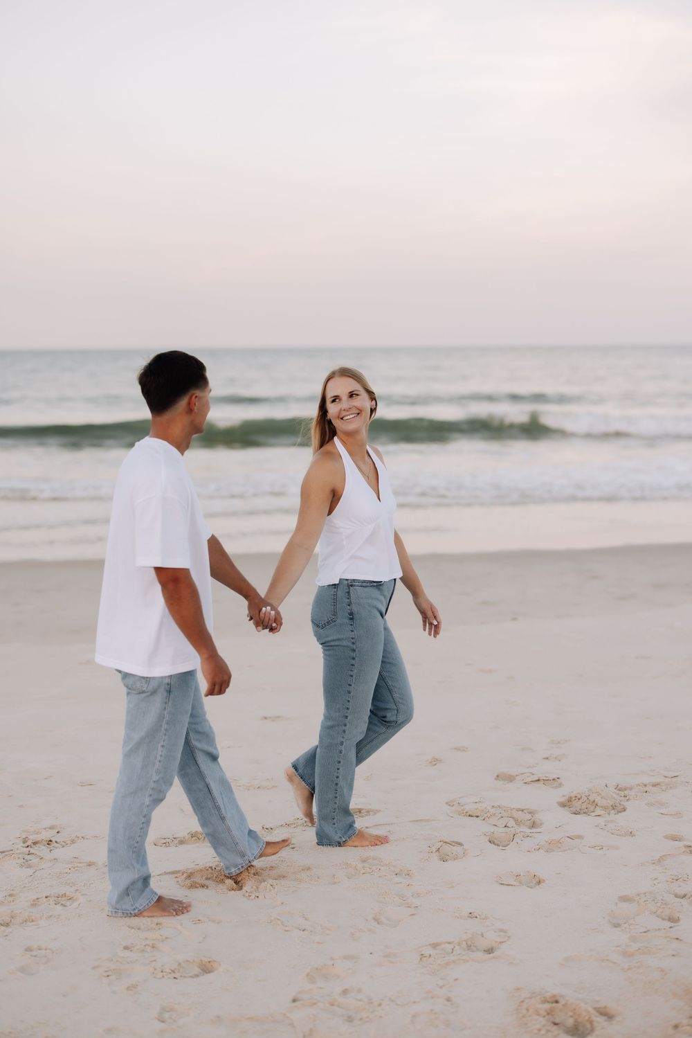 A couple holding hands and walking barefoot on a sandy beach at sunset, both wearing white tops and jeans.
