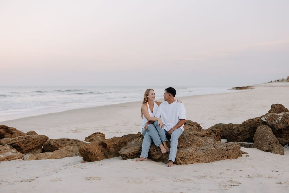Couple sitting on rocks at a sandy beach during sunset, smiling at each other with ocean waves in the background.