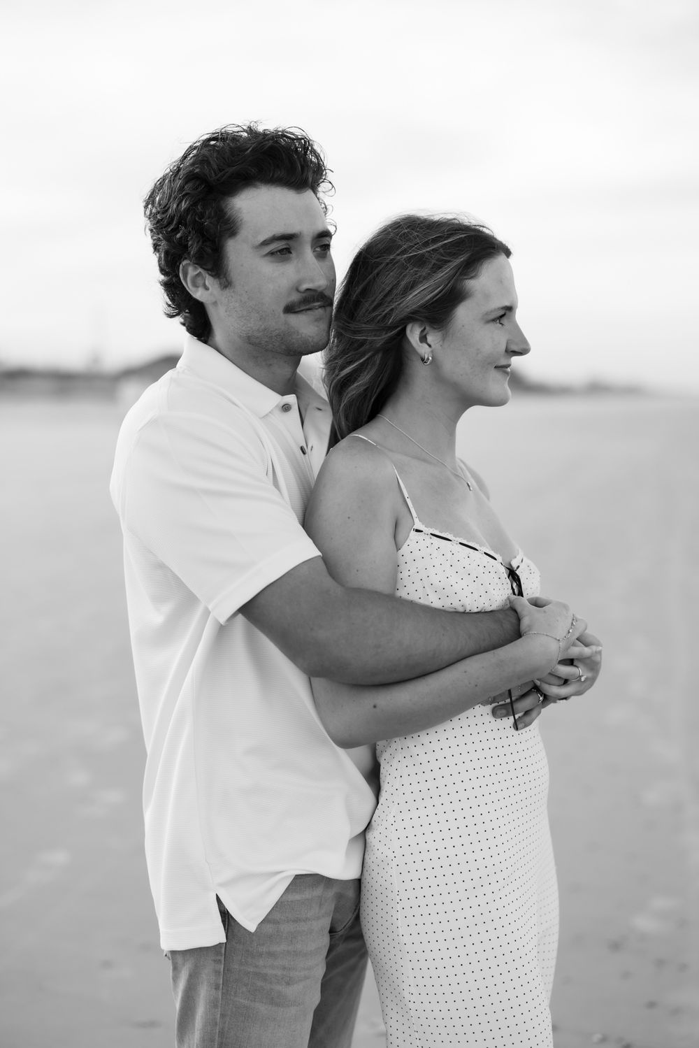 Black and white photo of a couple embracing on a beach, man hugging woman from behind as they gaze into the distance.