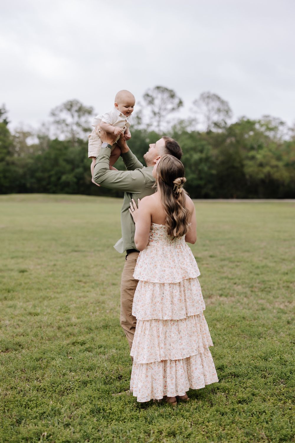 A joyful family outdoors, with a man lifting a baby while a woman in a floral tiered dress looks up smiling.