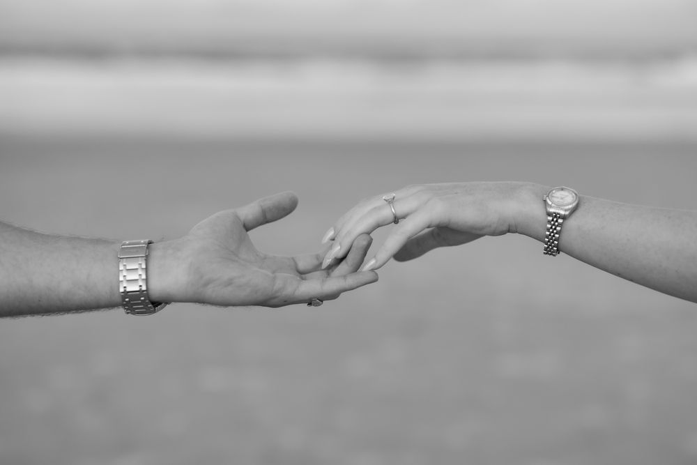Black and white photo of two hands gently touching, each wearing a watch, with a blurred beach background.