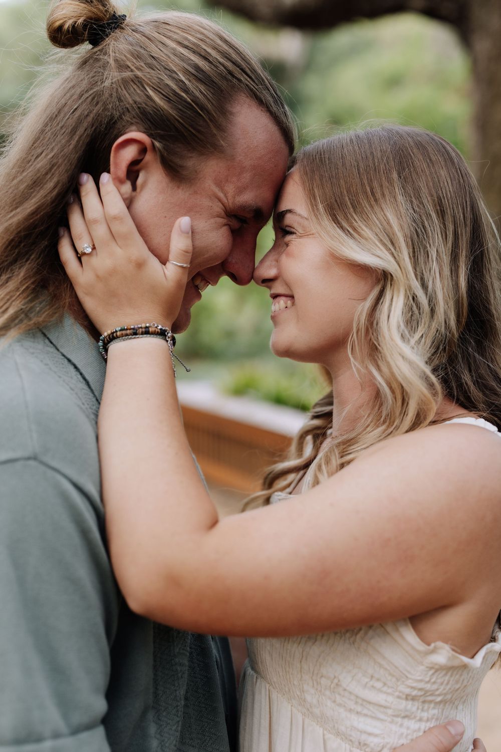 A couple shares a romantic forehead touch, smiling closely, with the woman wearing an engagement ring outdoors.