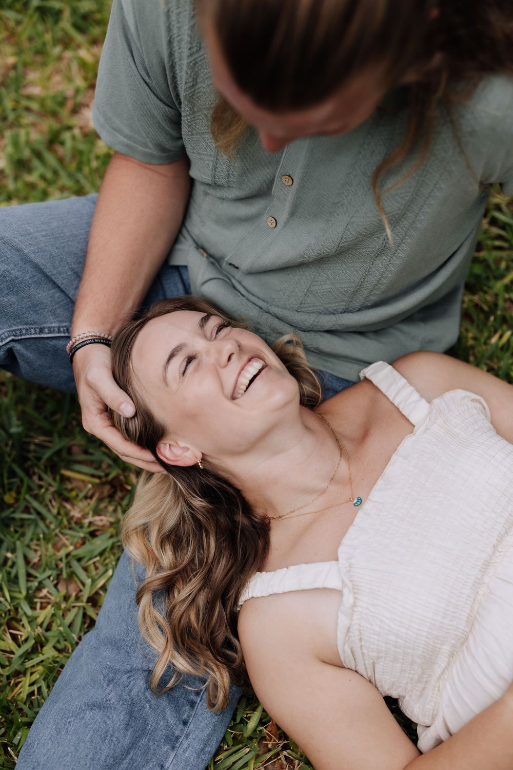 A woman laughs joyfully while lying on grass as another person gently touches her hair outdoors.