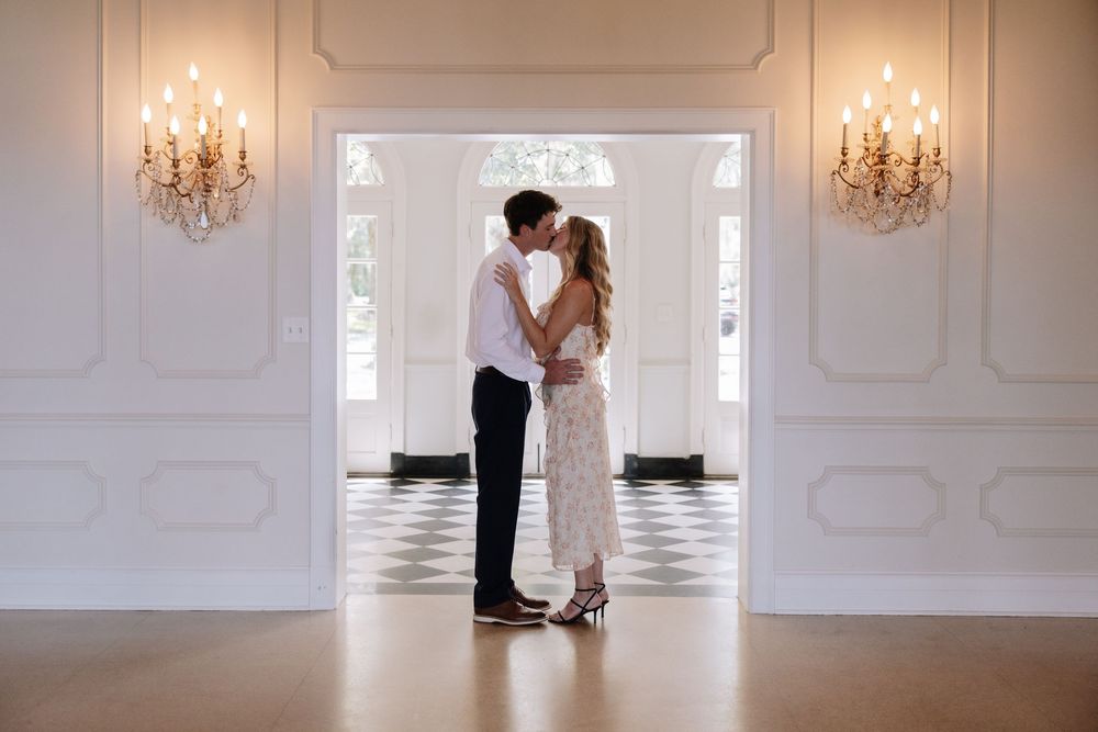 Couple sharing a romantic kiss in an elegant white ballroom with ornate chandeliers and checkered floors.