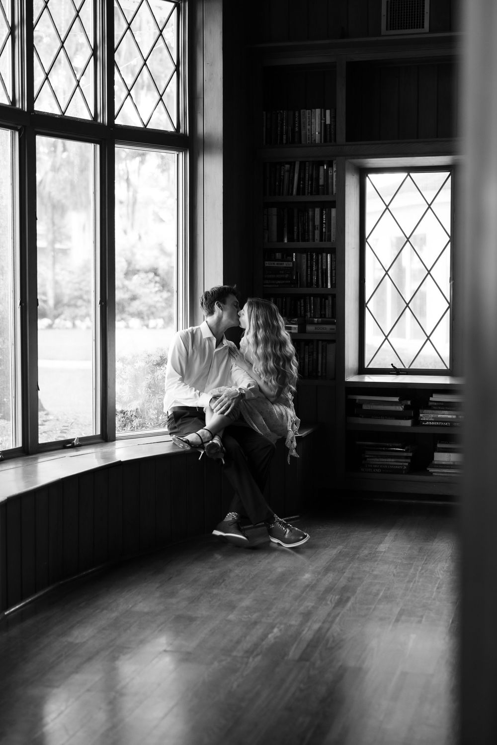 Black and white photo of a romantic couple sitting together on a window seat in a cozy library with bookshelves.