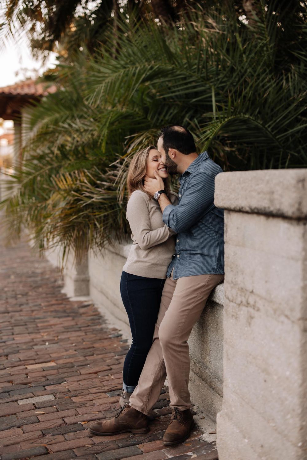 A couple shares an intimate moment on a brick walkway surrounded by palm trees and stone walls.
