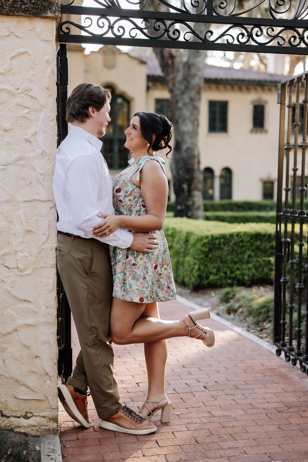 Couple embracing and smiling at each other by ornate iron gate, woman in floral dress, man in white shirt.