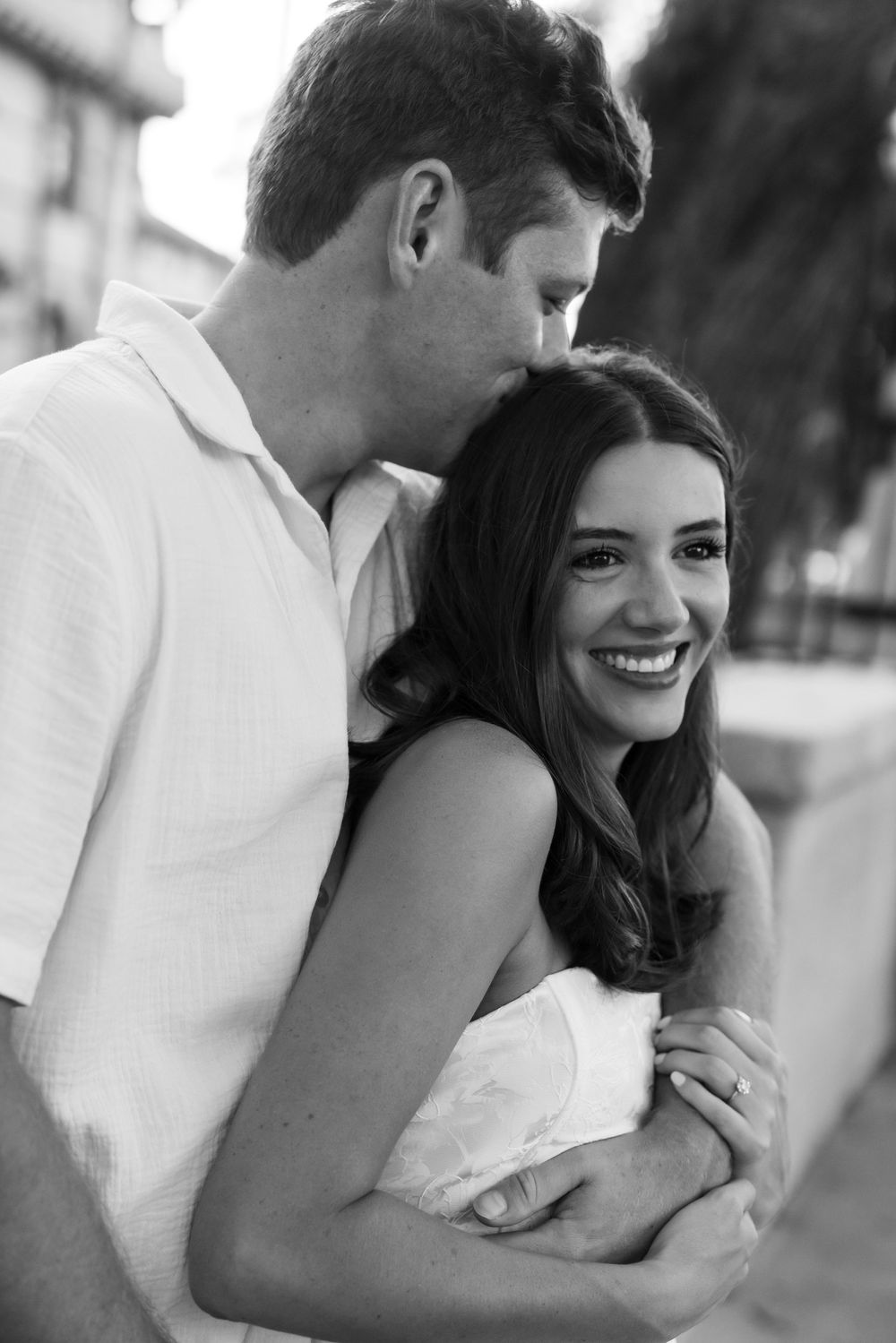 Black and white photo of a smiling couple embracing outdoors, man kissing woman's head while she laughs joyfully.