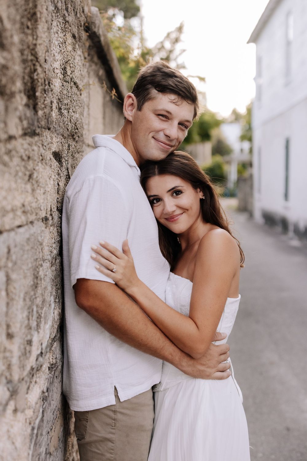A young couple in white outfits embracing against a stone wall on a charming street, smiling warmly at the camera.