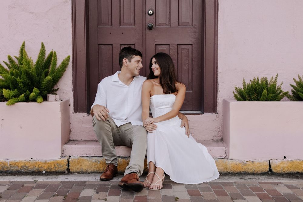 Couple sitting on steps in front of a pink building with a dark door, smiling at each other, wearing white outfits.
