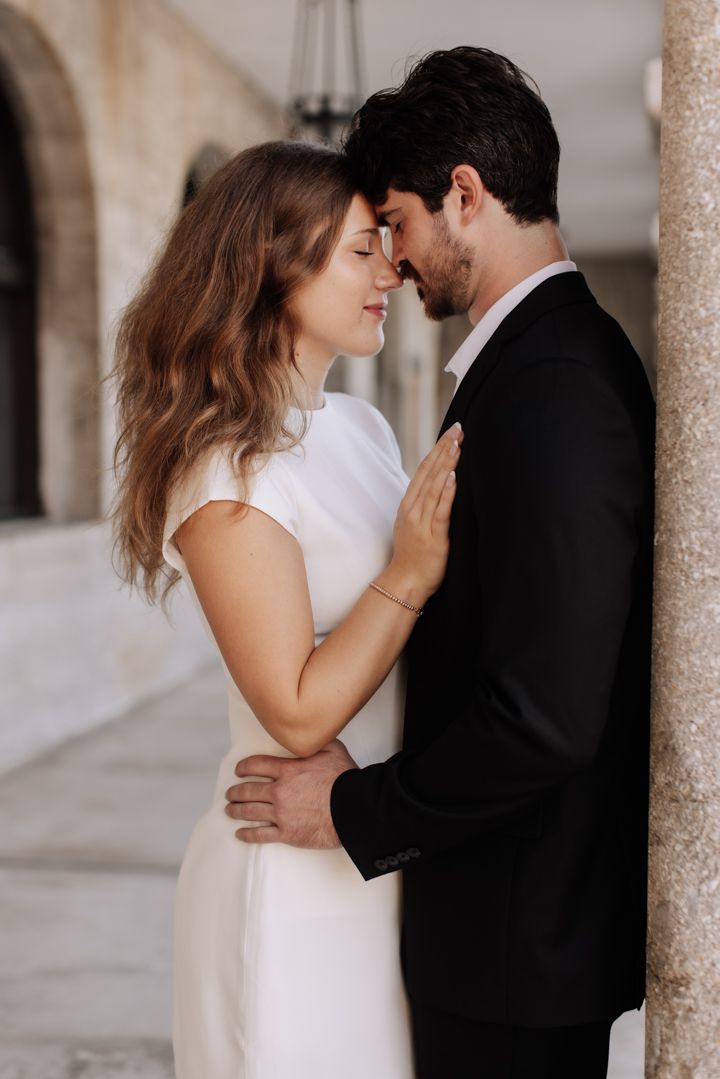 A romantic couple in wedding attire share an intimate moment against a stone column backdrop.