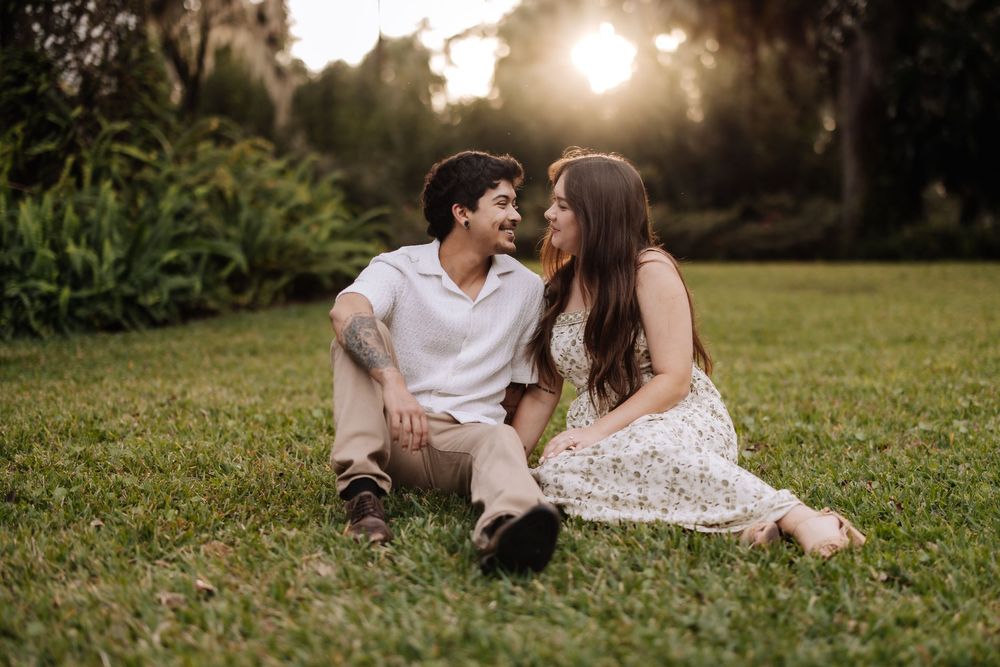 A couple sits together on grass during golden hour sunset in a romantic outdoor setting.