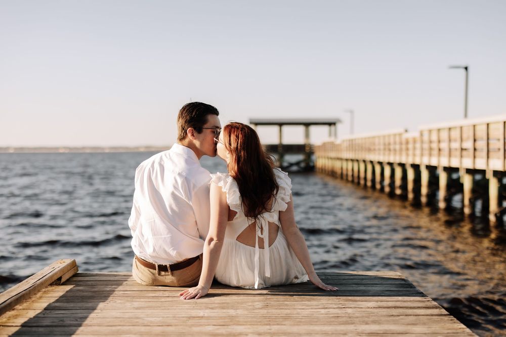 A couple sits together on a wooden pier at sunset overlooking the ocean in romantic silhouette.