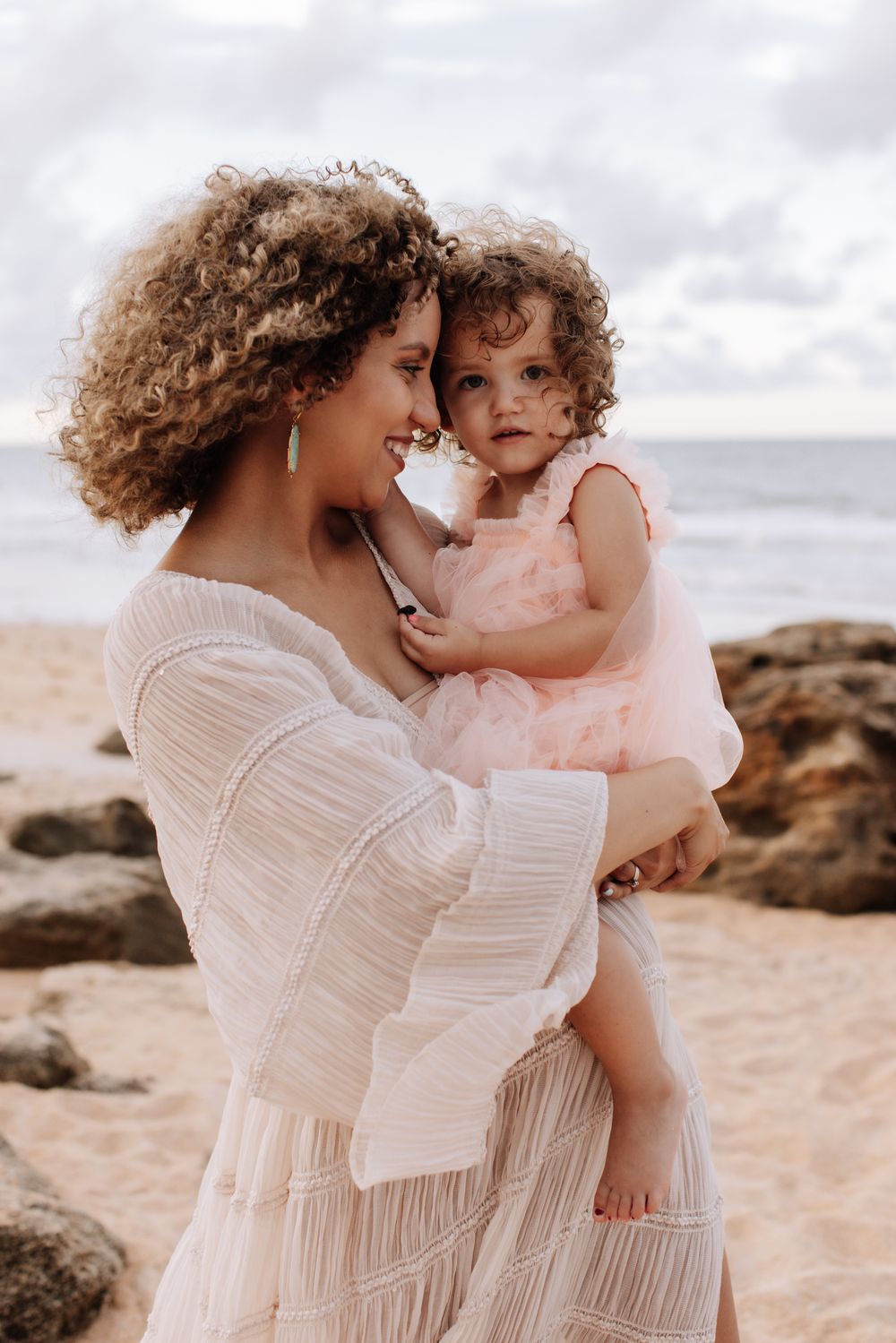 A curly-haired mother in a white dress holds her toddler daughter in a pink tutu on a sandy beach.