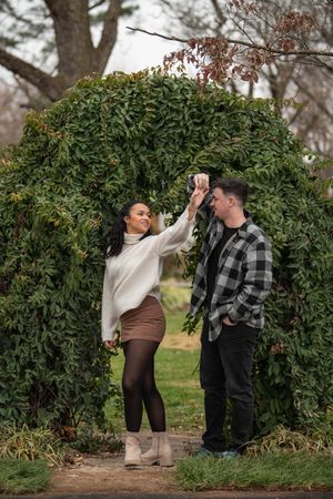 A couple shares playful moments near a green hedge in a park setting.
